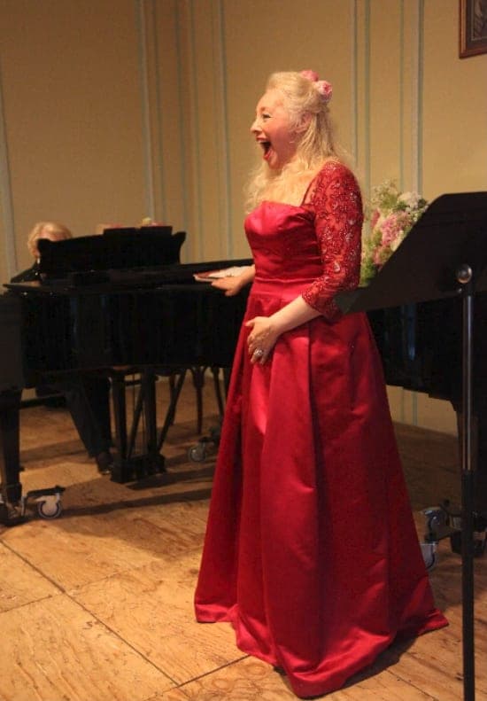 Melissa en robe rouge, chantant en récital, un piano derrière elle
